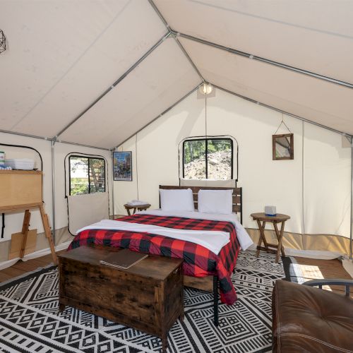 A cozy tent bedroom with a bed, plaid blanket, wooden chest, side tables, a sofa, rug, and vintage-look dresser; bright windows inside.