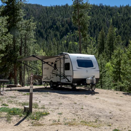 A white travel trailer parked on a dirt clearing amid a forested campground, with a wooden picnic table and trees surrounding it.