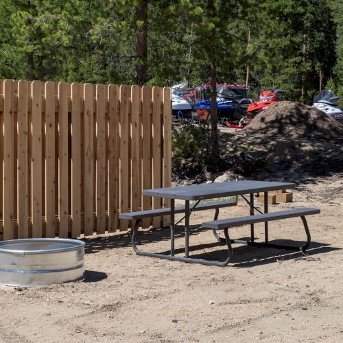 A wooden privacy fence, a metal picnic table, a round metal fire ring, and dirt ground with trees and parked cars in the background.