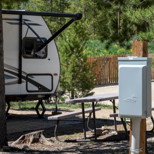 A parked RV with an awning extended, near a picnic table and a utility pedestal, in a wooded campsite.