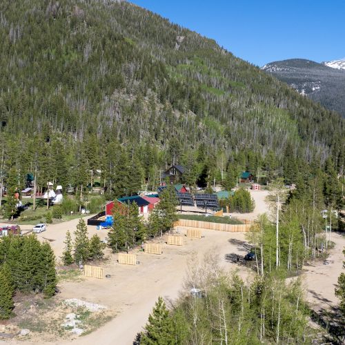 A small mountain town with colorful cabins, a dirt road grid, and a forested hillside backdrop under a blue sky.