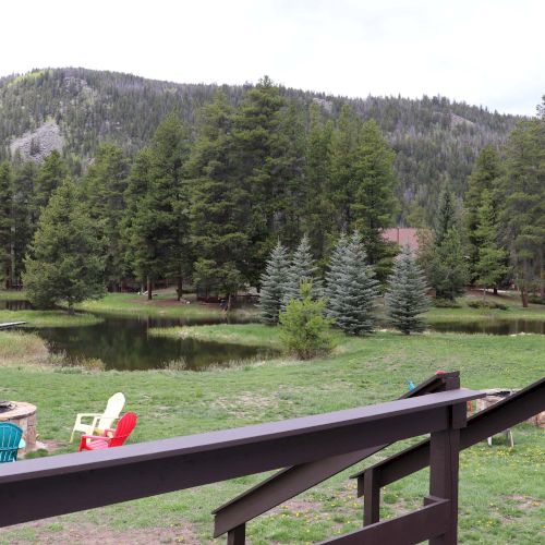 A grassy yard with a small pond, pine trees, and mountains in the background; a wooden fence and colorful chairs sit by the water.