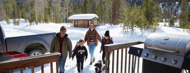 A family is outside in the snow with a dog, near a grill and picnic table, set in a wintery hillside yard.