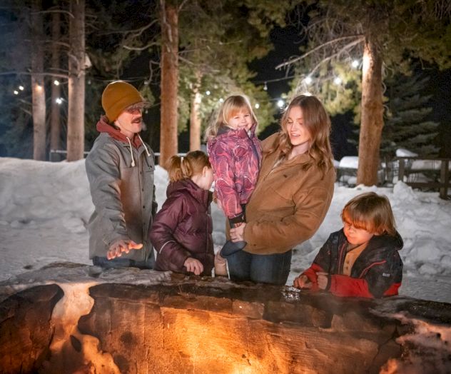 A group of people sitting around a campfire, smiling, sharing a cozy outdoor moment.