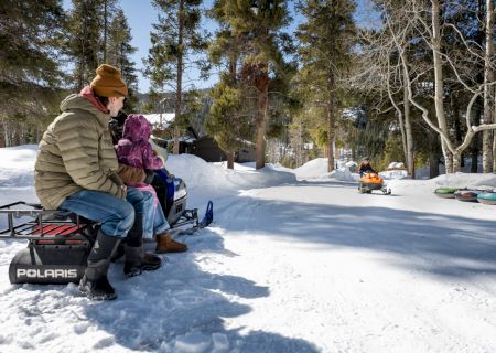 A person wearing winter gear sits on a snowy slope, with trees and a wooden cabin in the background.