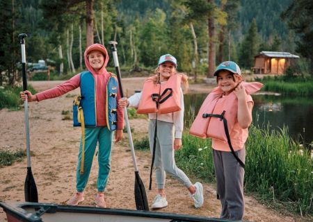 Three kids stand on a sandy lake shore with paddles and life jackets, ready for a boat outing as trees and cabins glow in the background.