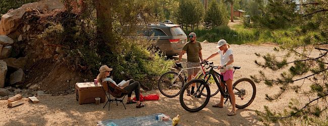 People camping in a forested area: a couple sits on chairs, gear laid out on a blanket, and two children with bikes near a parked SUV.