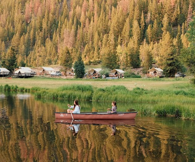 Two people in a red canoe on a calm lake, with a forested mountainside and a small riverside town in the background.