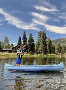 A person stands on a blue canoe, fishing on a calm lake surrounded by pine trees, mountains, and a few lakeside cabins under a blue sky.
