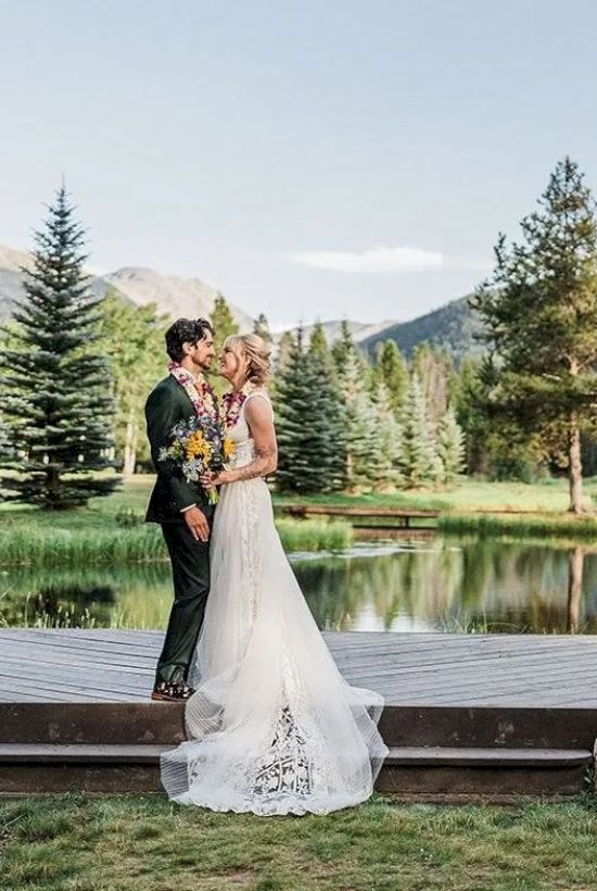 A couple in wedding attire stands on a wooden platform by a pond, surrounded by green meadow and tall trees in a mountain setting.