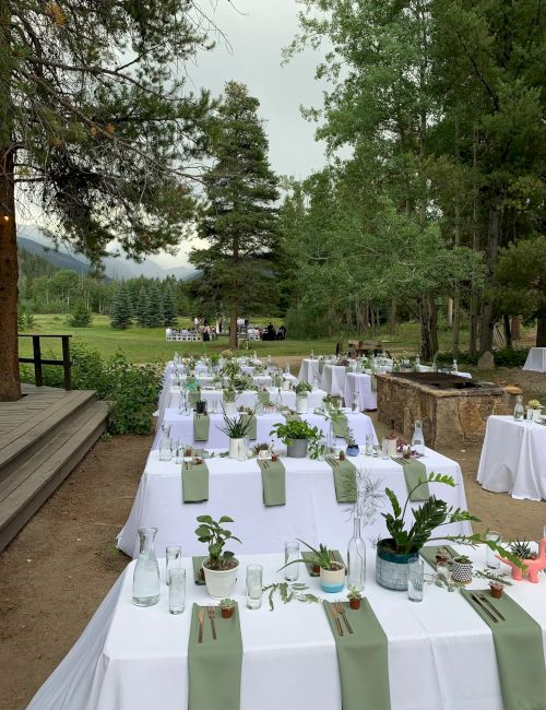 An outdoor wedding reception setup with long tables, white linens, green runners, potted plants, and lanterns in a forested yard.
