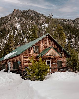 A rustic log cabin with a green metal roof sits in a snowy mountain landscape, pine trees surrounding it under a cloudy sky.