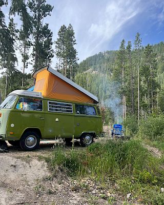 A green vintage van with a raised orange pop-top sits by a forested campsite; a small blue tent or vehicle in the background and smoke rising gently.