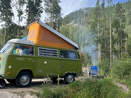 A green vintage van with a raised orange pop-top sits by a forested campsite; a small blue tent or vehicle in the background and smoke rising gently.