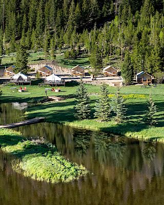 A tranquil lakeside village with wooden cabins lining the shore, evergreen trees in the background, and a calm reflective water body nearby.