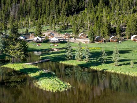 A tranquil lakeside village with wooden cabins lining the shore, evergreen trees in the background, and a calm reflective water body nearby.