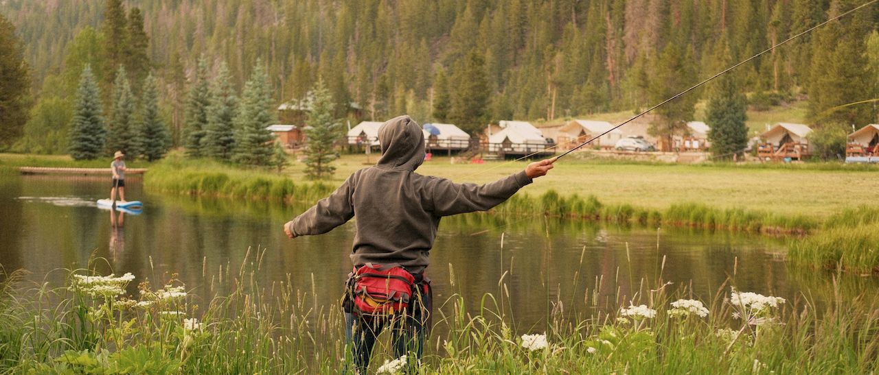 Two people fishing by a pond in a grassy lakeside with trees and cabins in the background; one toward the water, arms outstretched.
