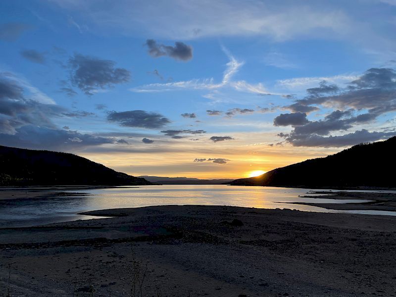 A tranquil beach at sunset with calm waters, dark silhouettes of hills, and a colorful sky streaked with clouds as the sun dips below the horizon.