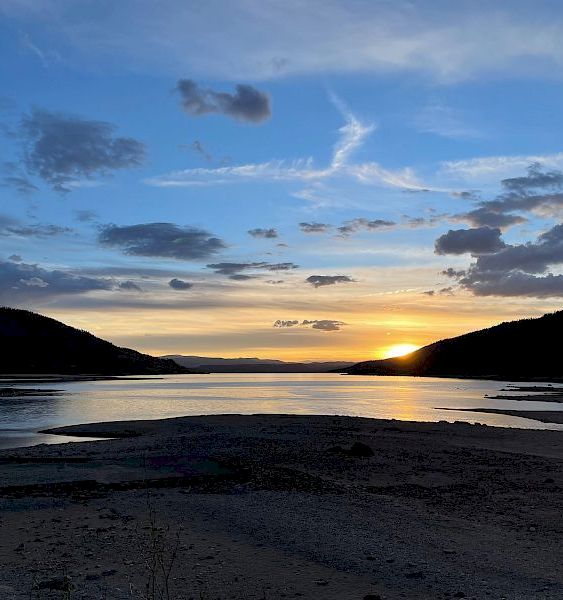 A tranquil beach at sunset with calm waters, dark silhouettes of hills, and a colorful sky streaked with clouds as the sun dips below the horizon.