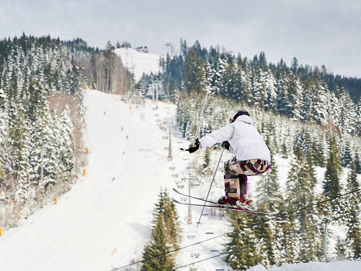 A skier in white gear glides down a snowy, forested slope, trees dusted with fresh snow under a cloudy sky.