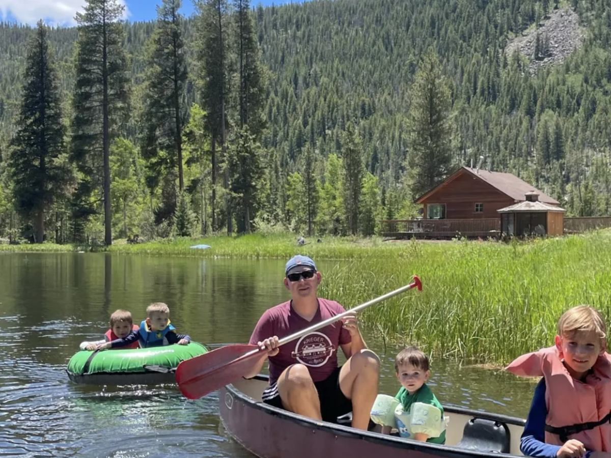 Four people, including two kids, canoe on a calm lake with one adult paddling in a red canoe while two kids ride in an adjacent green inner tube behind them. End of sentence.