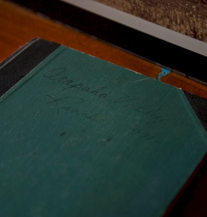A worn green notebook with handwritten text rests on a dark desk near a monitor; it looks old and slightly scratched, closed.