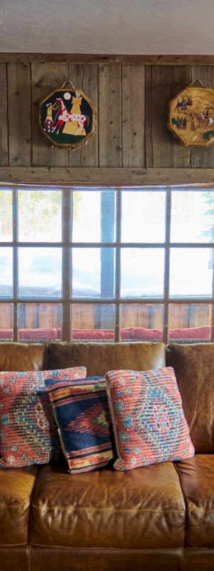 A cozy rustic living room with a brown leather sofa, wooden panel walls, a large window, decorative plates, a mounted deer head, and a bookshelf along the right wall.