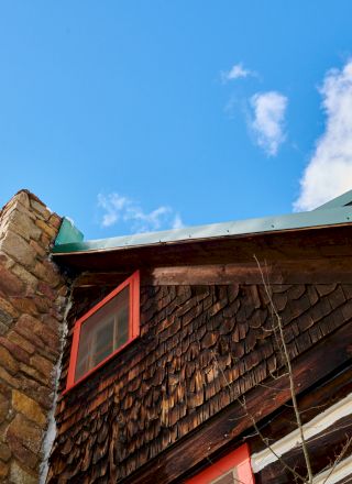 A rustic wooden cabin with a stone chimney, red-trimmed windows, and a slanted roof against a bright blue sky with a few clouds.