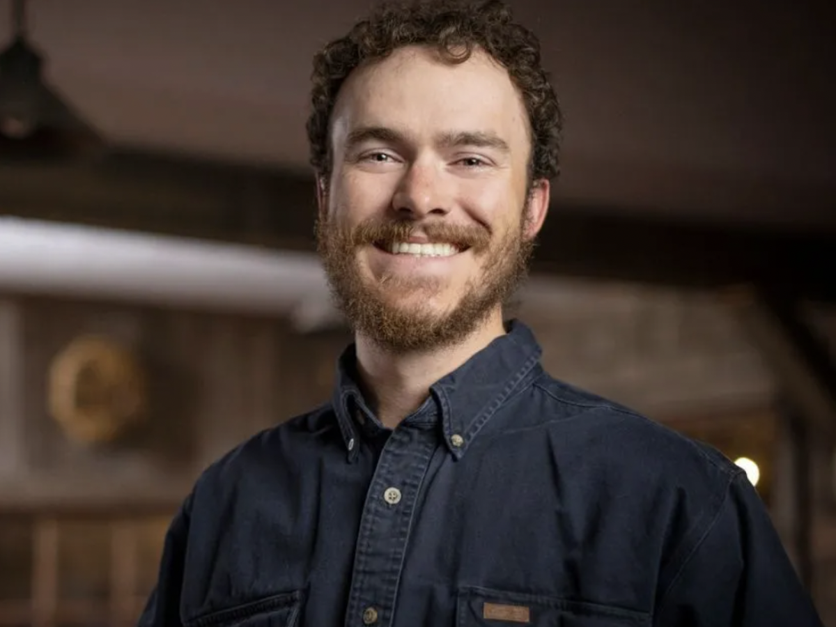 A smiling man with a beard wearing a dark blue button-up shirt, standing indoors with a warm, wooden background.