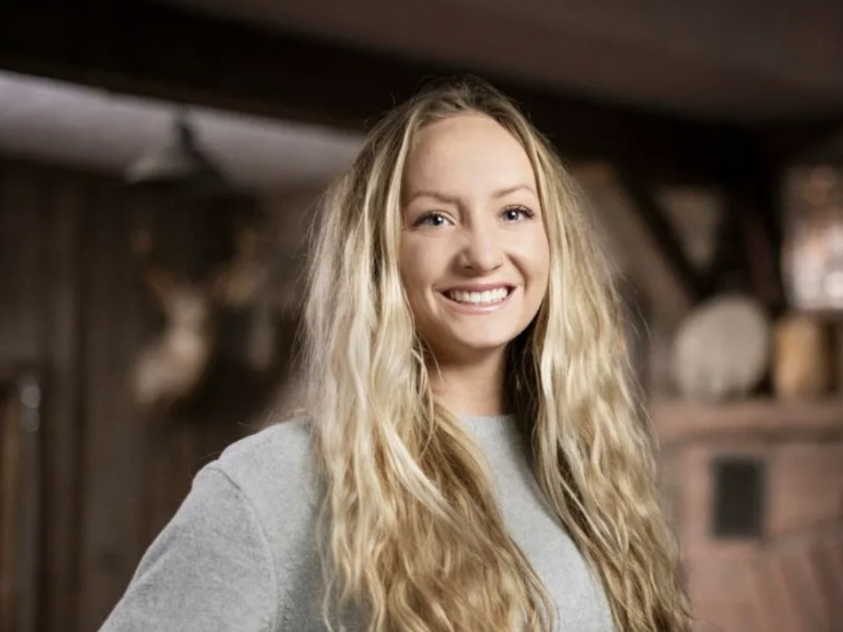 A smiling woman with long blonde hair wearing a light gray shirt, standing in a rustic wooden setting.