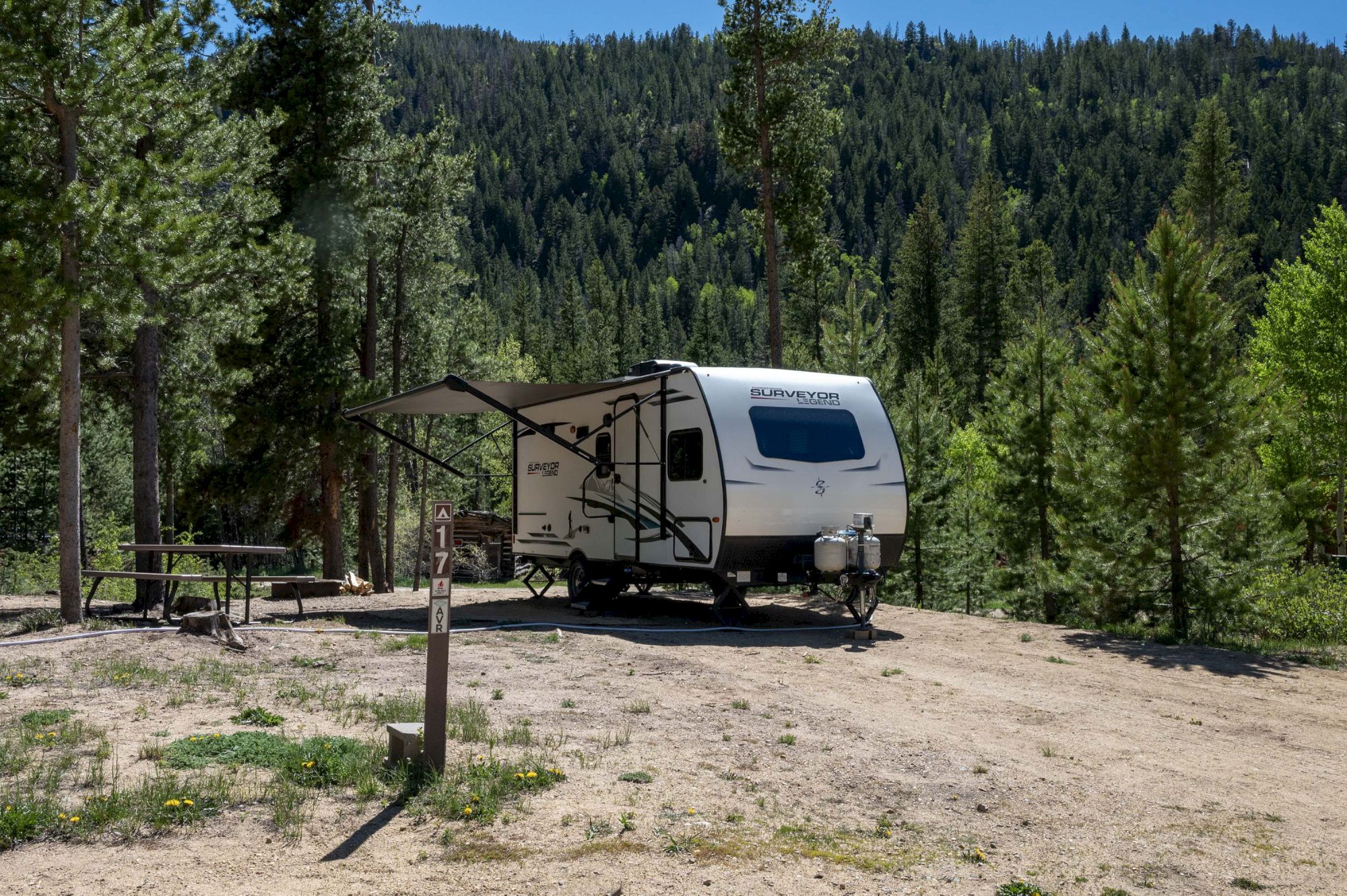 A small white travel trailer parked in a forest clearing, with a shaded awning, picnic table, and pine trees surrounding a sunny campsite.