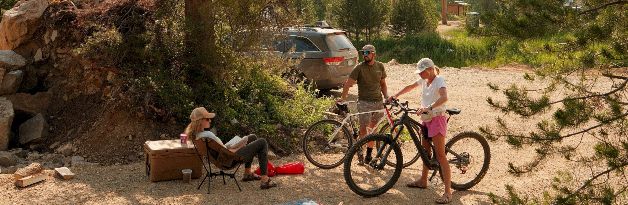 People relaxing outdoors by a parked SUV and bikes; a makeshift campsite with a tarp, cooler, and chairs under trees in a forested area.