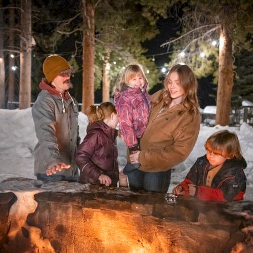 A family gathers around a glowing fire pit at night in a snowy outdoor setting, kids bundled up, chatting and smiling together.