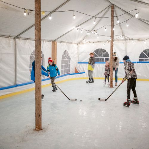 People skating on an indoor rink under string lights, kids and adults with hockey sticks, small wooden posts, white tent walls.