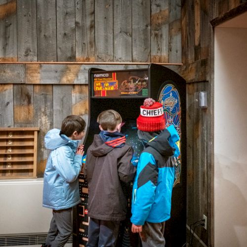 Kids gathered around a vintage arcade cabinet in a cozy, wood-paneled room, wearing jackets and hats, exploring a retro gaming machine.