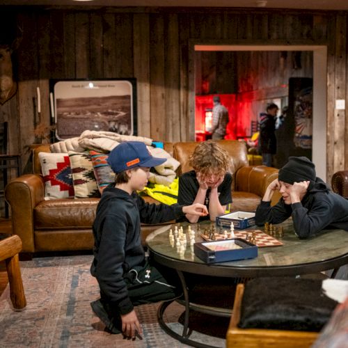 Three kids sit around a glass table playing a board game in a cozy, wood-paneled room filled with couches and vintage decor, focused on the game.