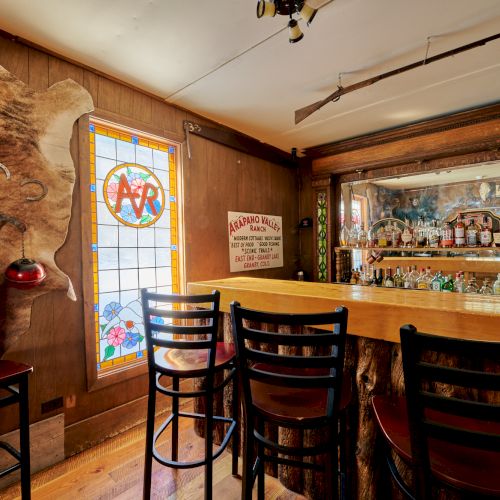 A cozy rustic bar with wooden walls, a paneled back bar, bottles on shelves, a stained glass door, and four tall wooden stools at the counter.