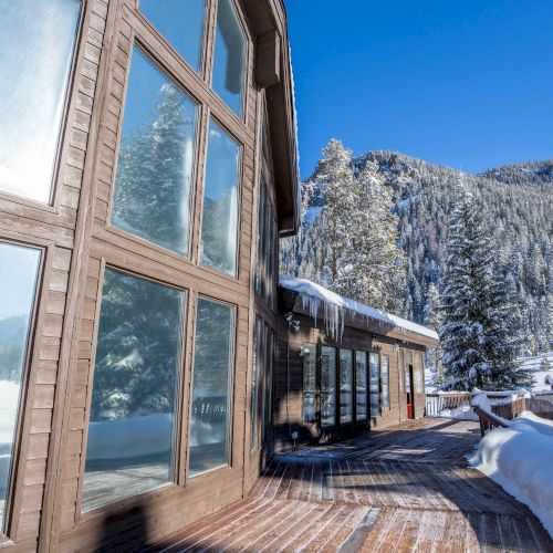 A wooden chalet beside a snow-covered path and tall pine trees, with clear blue sky and mountain backdrop.