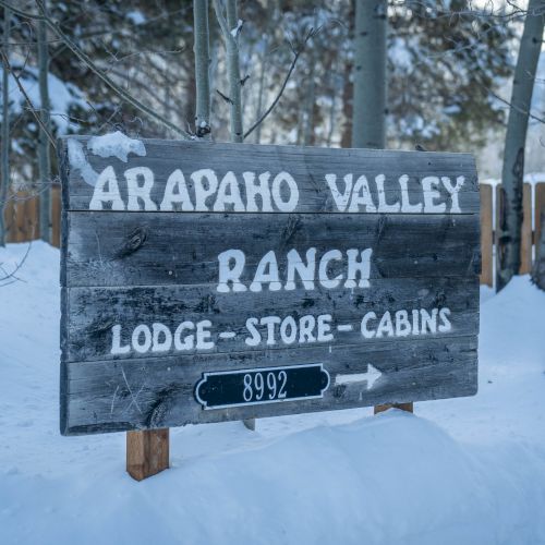 A weathered wooden sign reads &ldquo;ARAPAHO VALLEY RANCH&rdquo; with &ldquo;LODGE &ndash; STORE &ndash; CABINS&rdquo; and a small address plaque in snow, pointing right.