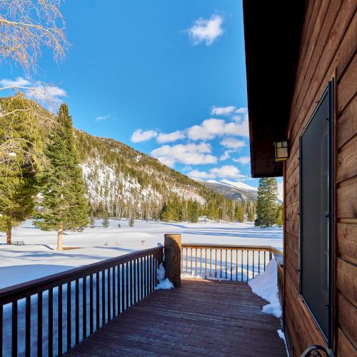 Snowy deck overlook of a wooden cabin with a frosty lake, pine trees, and distant mountains under a bright blue sky.
