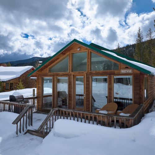 A cozy wooden cabin with large glass windows sits in fresh snow, a wraparound deck with lounge chairs; cloudy skies loom above the pine trees.