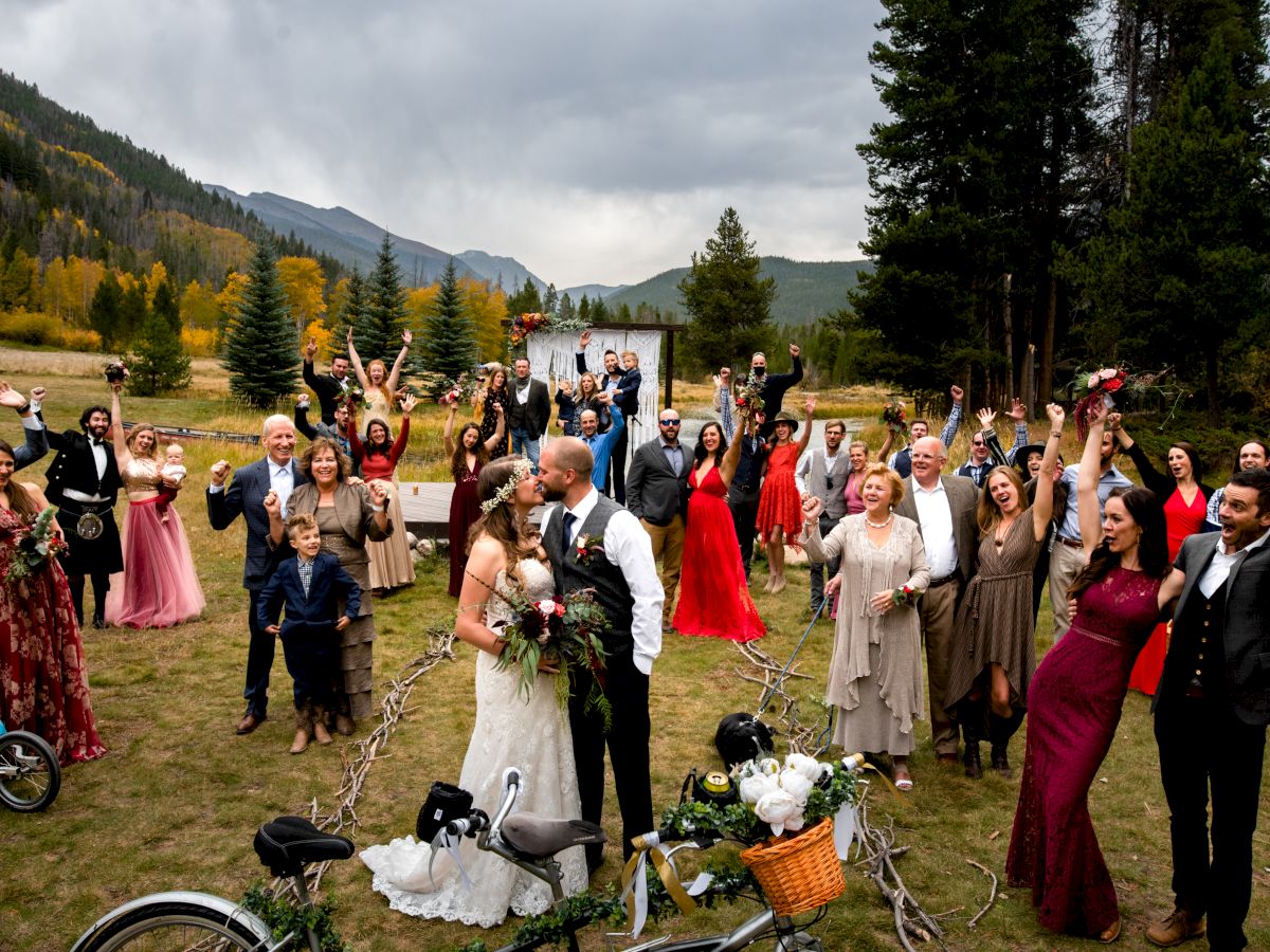 A wedding party cheers in a mountain meadow, bride and groom at center kissing, guests in formal attire raising hands, bikes and baskets nearby.