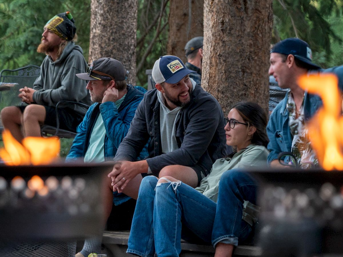 A group of friends sits around a fire pit in a wooded area, chatting and relaxing near the flames.
