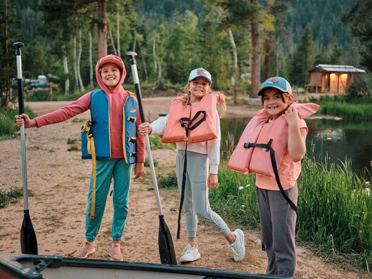 Three kids in life jackets stand with paddles on a lakeshore, ready to kayak, smiling near a boat and a cabin by the water.