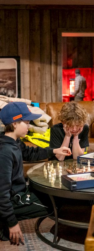 Three kids play a round table board game in a cozy, cluttered lounge with couches, a mirror, and warm wood paneling in the background.