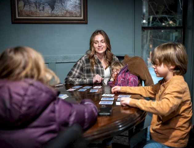A group of people sits around a table, snacks and drinks visible, as they chat in a warmly lit living room.