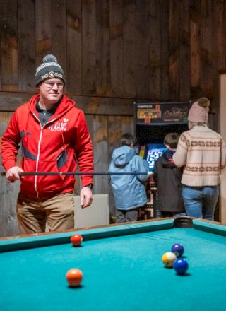 Two people play pool in a cozy bar; balls scatter near the table while one player lines up a shot, and a warm, rustic setting surrounds them.