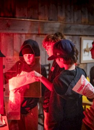 A group of people in a dim, red-lit setting, possibly a party or nightclub, with one person in the foreground wearing a red jacket and others mingling.