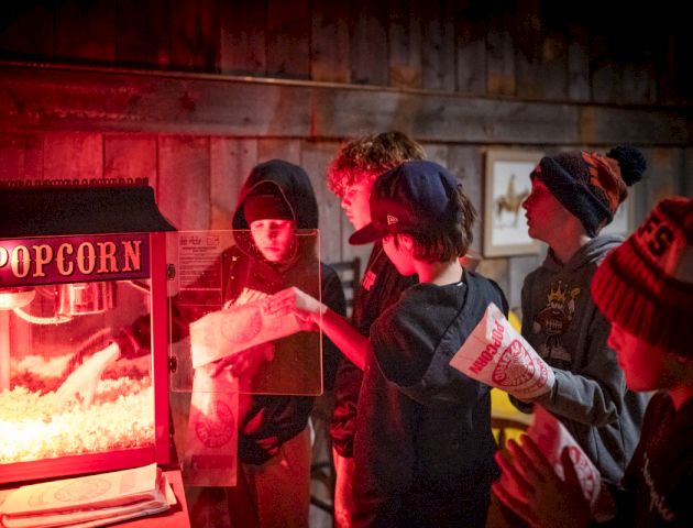 A group of people in a dim, red-lit setting, possibly a party or nightclub, with one person in the foreground wearing a red jacket and others mingling.