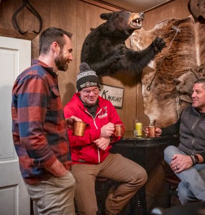 A group of five people sit and stand in a living room, smiling and posing for the camera, with a warm, casual gathering vibe.